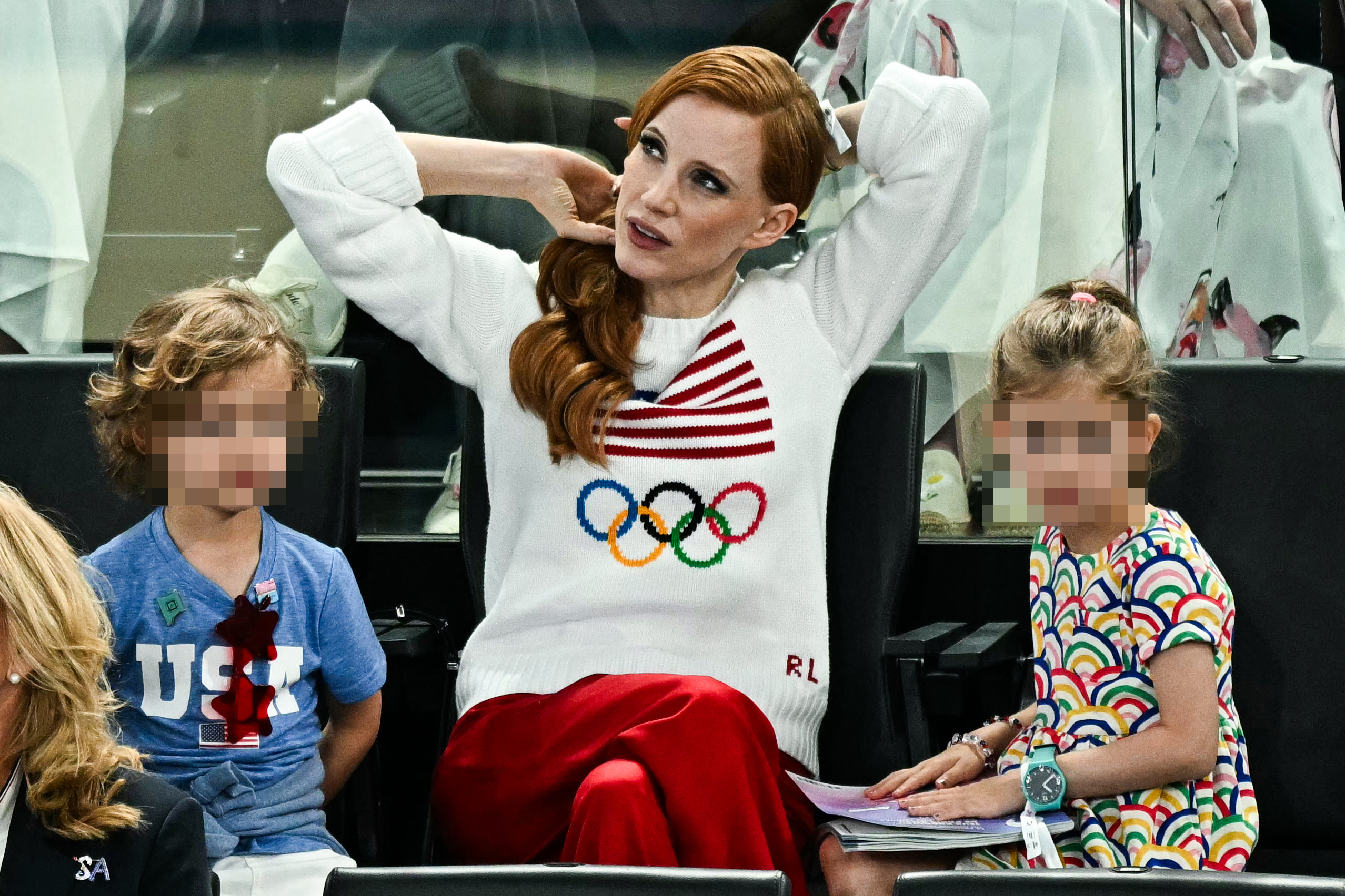 Jessica Chastain is sitting with two children at an event. She wears a sweater with the Olympic rings and a red and white design, paired with red pants