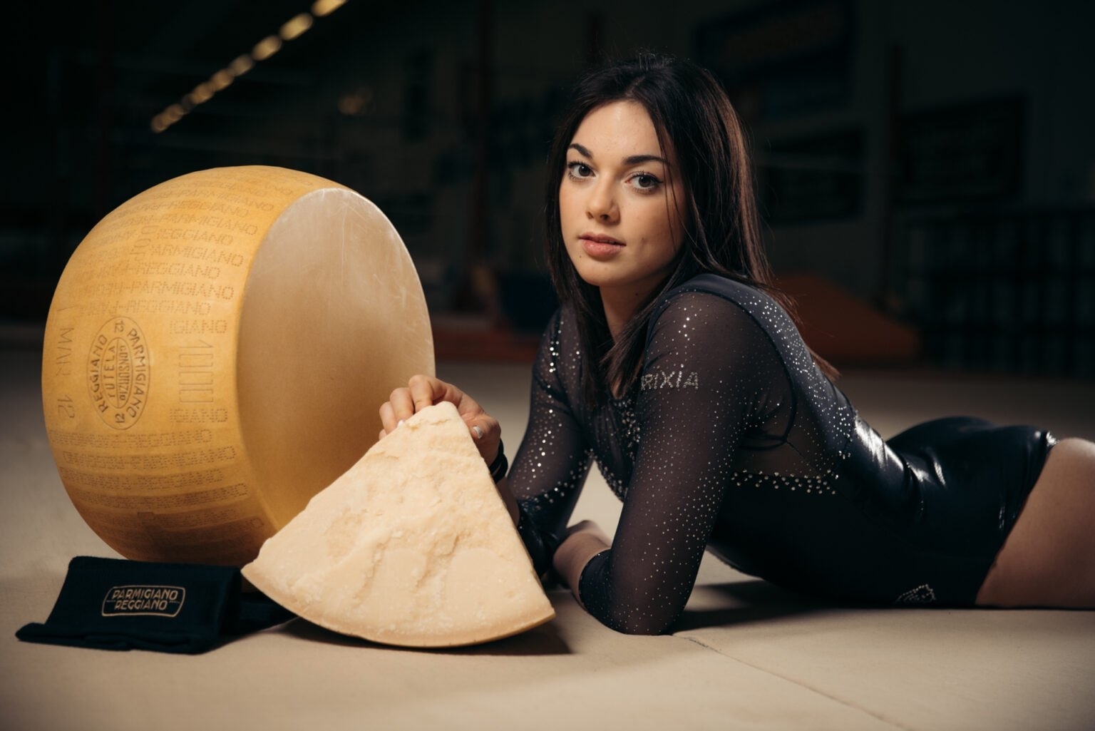 Olga Carmona, in a black gymnastic leotard, poses with a wheel and a wedge of Parmigiano-Reggiano cheese while leaning on the floor