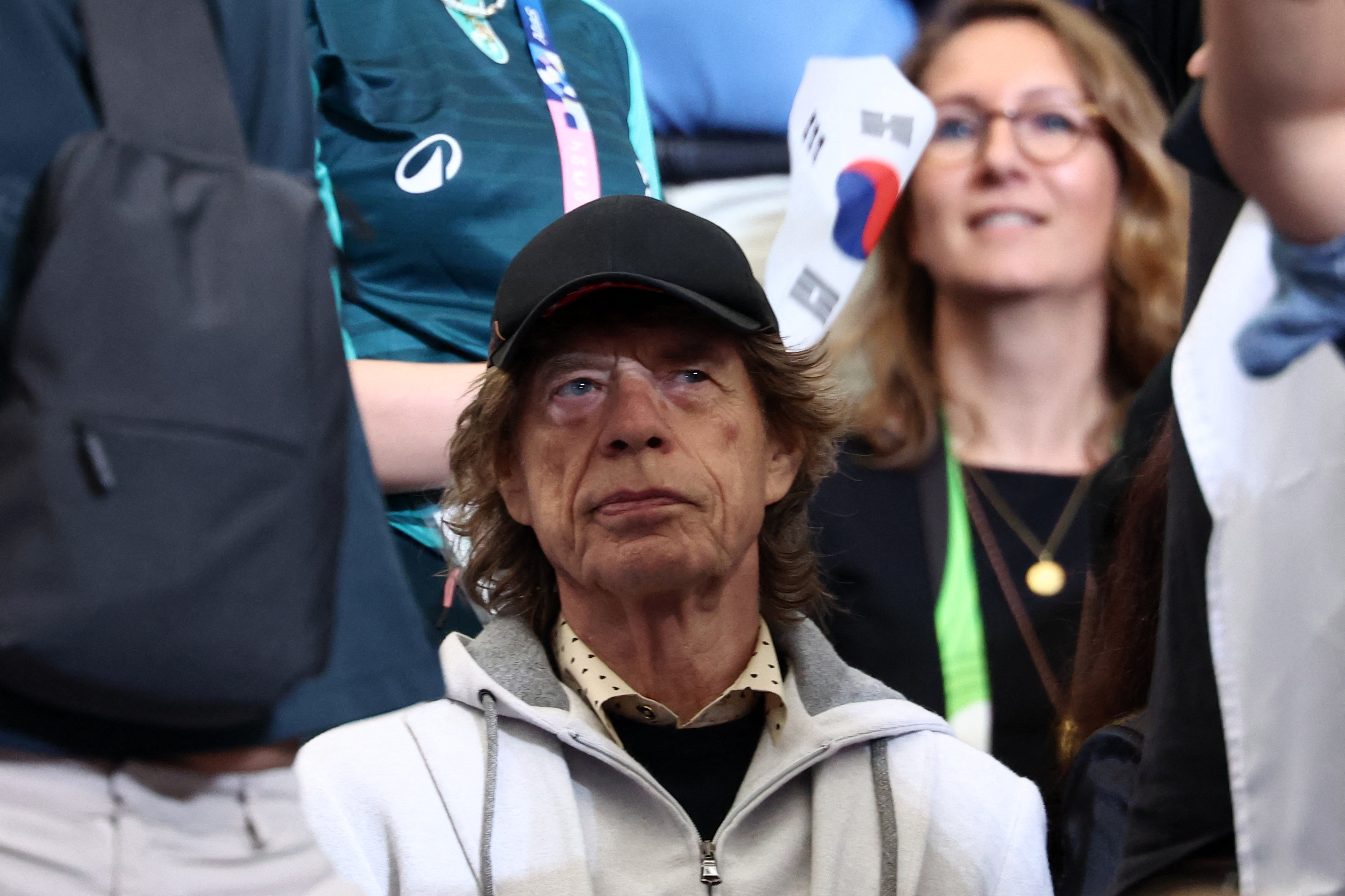 Mick Jagger stands among a crowd wearing a black cap and casual clothing. Faces around him appear focused on an event