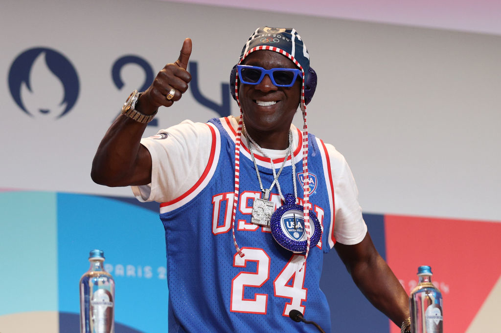 Flavor Flav, wearing a decorative headpiece, sunglasses, and a blue USA 24 jersey, gives a thumbs-up at an event