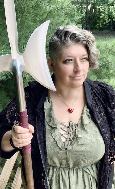 Person holding a large medieval-style halberd, wearing a lace-up dress and a heart pendant necklace, standing outdoors with trees in the background