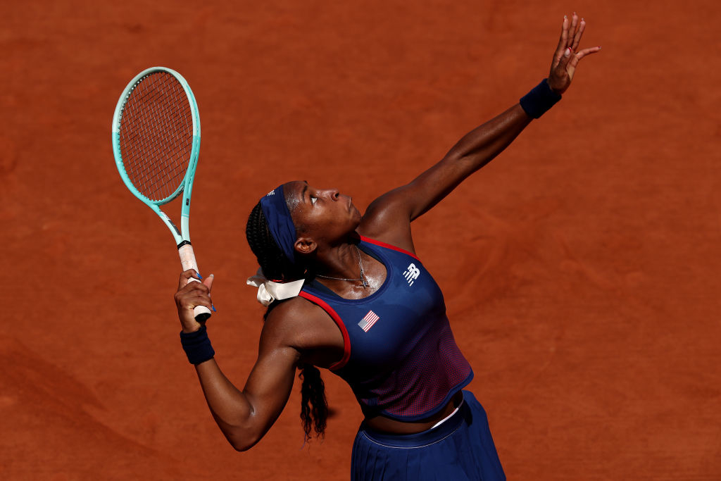 Coco Gauff prepares to serve during a tennis match on a clay court. She wears athletic gear, including a headband and wristbands