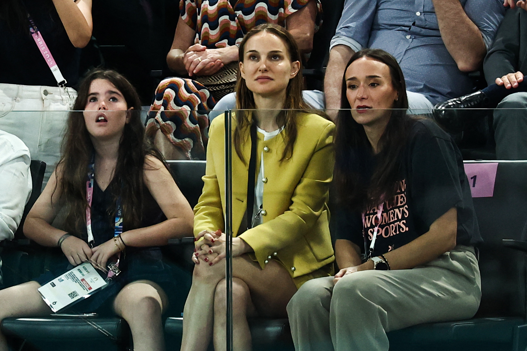 Natalie Portman sits with two women at a sports event, watching intently. One woman wears a Women's Sports t-shirt