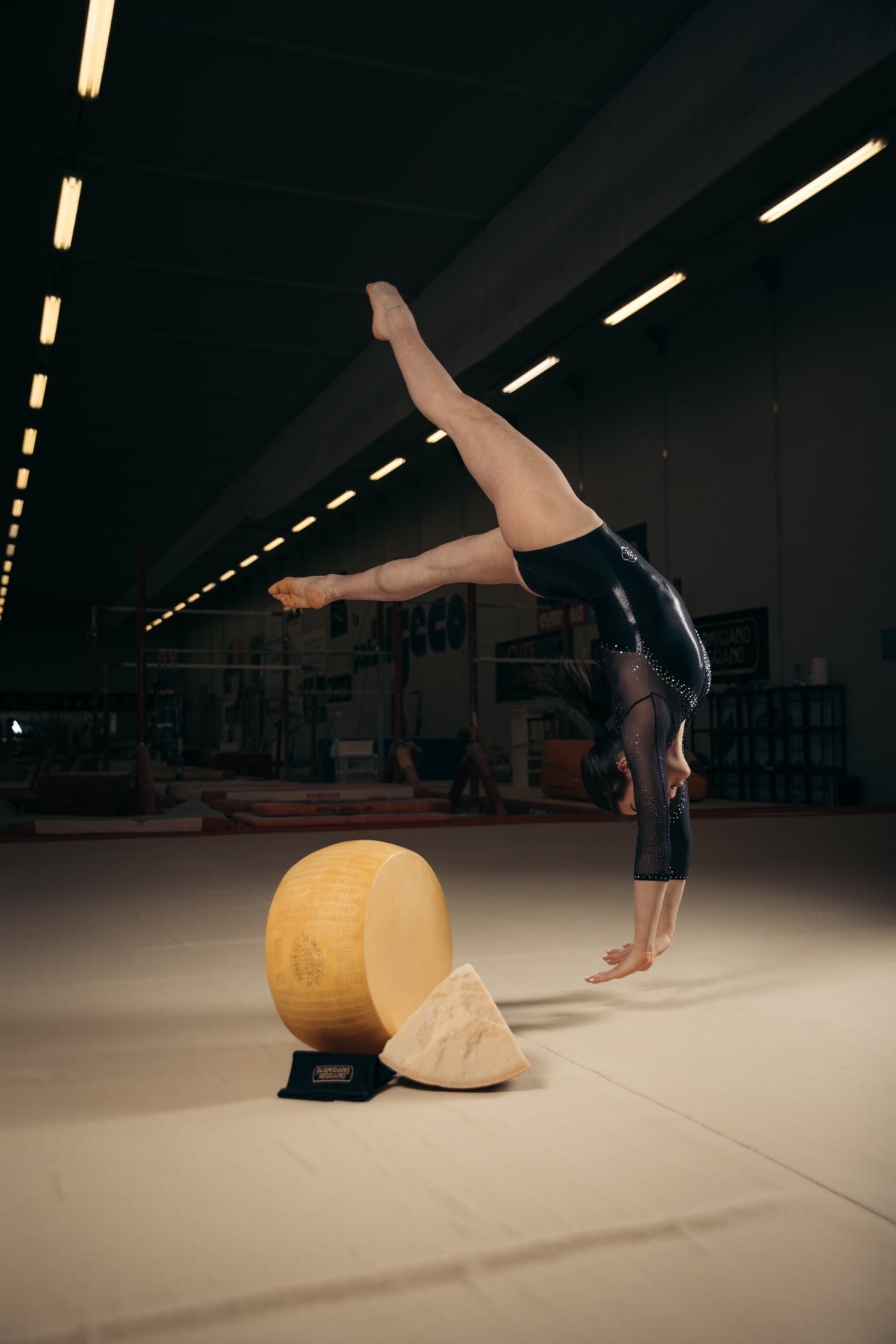 A gymnast performs a handstand in a gymnastics facility next to a large wheel of cheese and a cheese wedge on a black stand