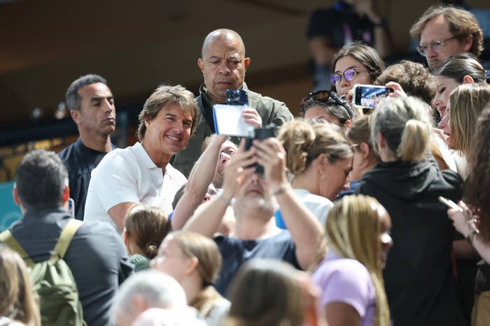 Tom Cruise smiles as he stands among fans taking selfies at a crowded event. He is wearing a white shirt