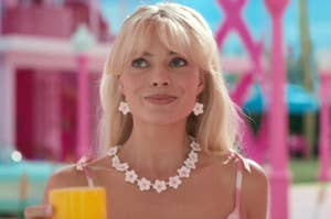 Margot Robbie wearing floral jewelry holds a cup, with a vibrant, playful background a Barbie