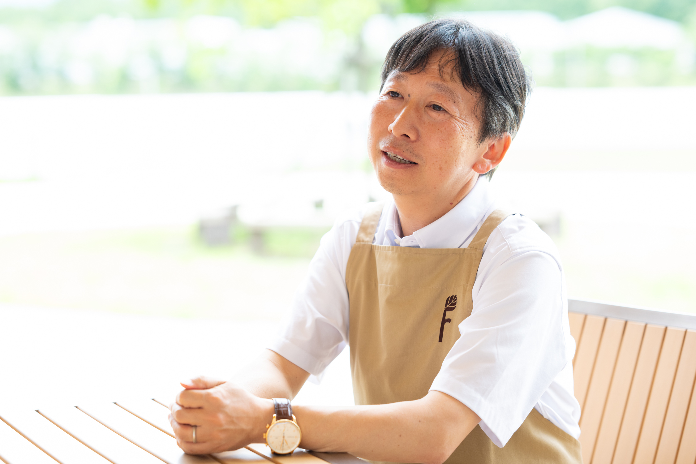 男性が淡い茶色のエプロンを着て、テーブルに座って話しています。背景は屋外の景色です。