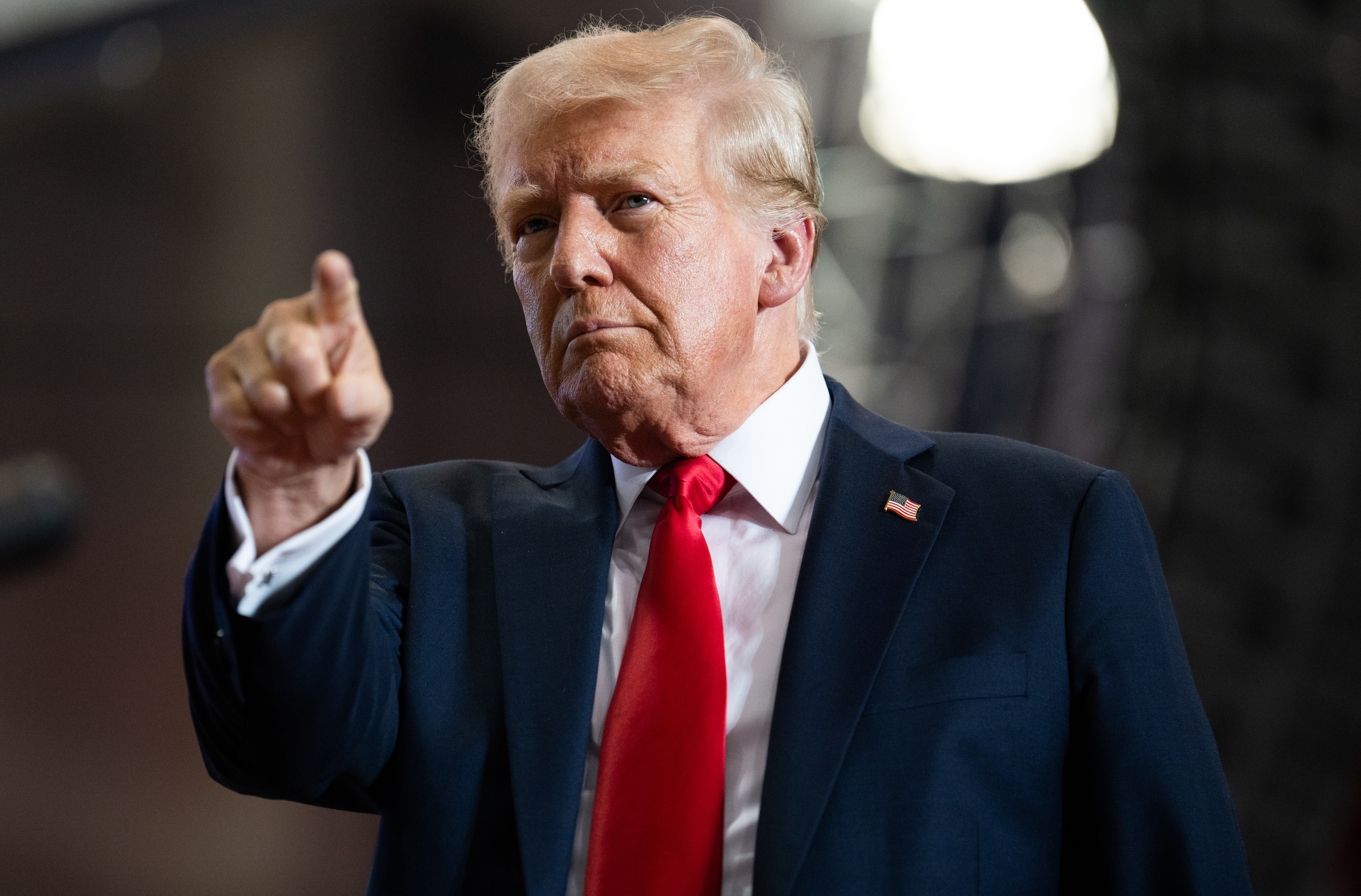 Donald Trump wearing a dark suit and red tie, pointing forward while speaking at an event