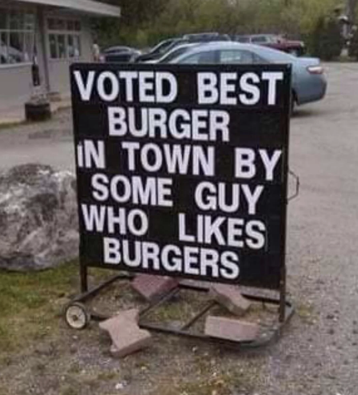 Sign reads: "Voted best burger in town by some guy who likes burgers," with a parking lot and cars in the background