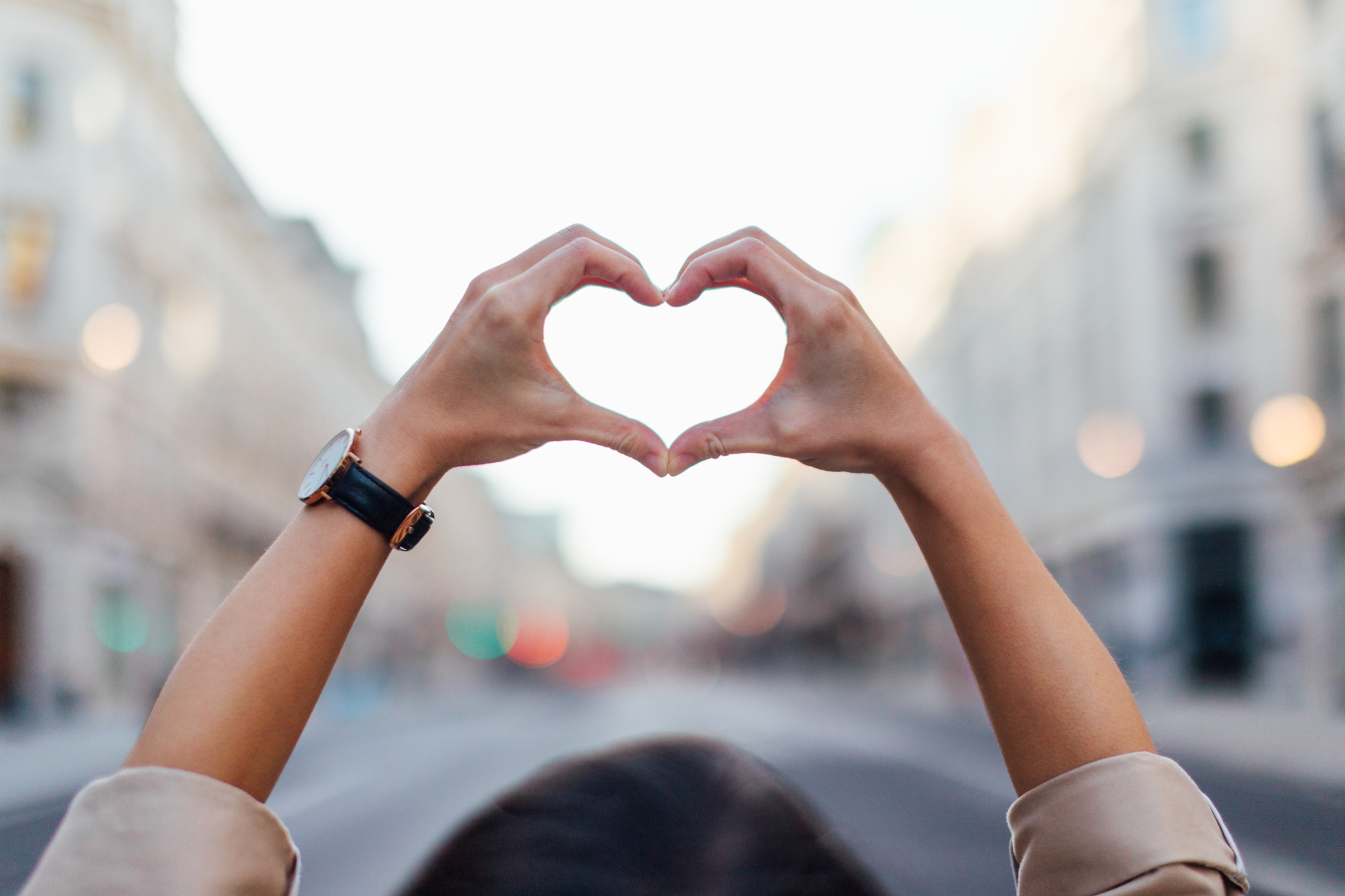 Person forming a heart shape with their hands, held up against an urban street backdrop