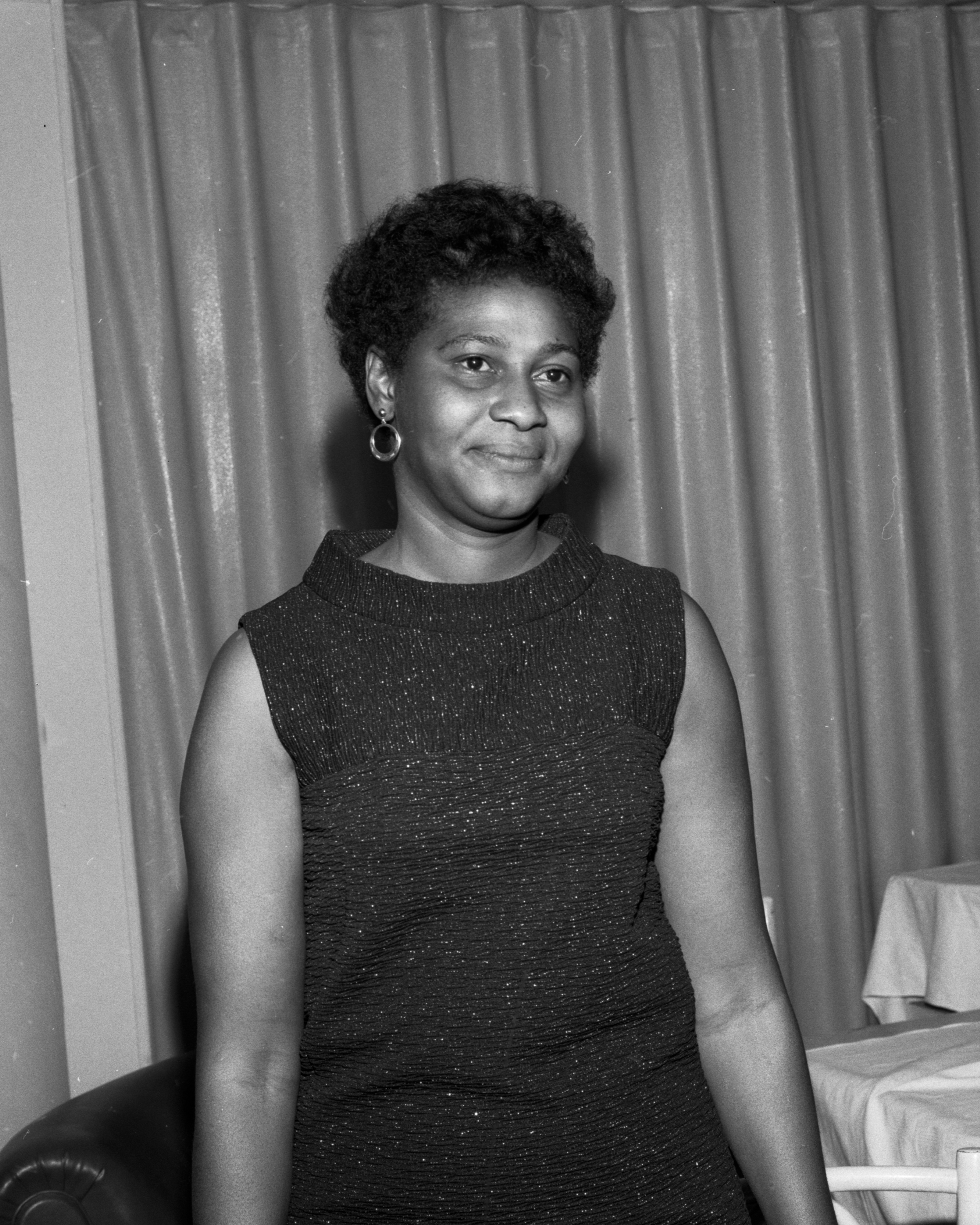 A woman wearing a sleeveless dress stands in front of a curtain, smiling at the camera