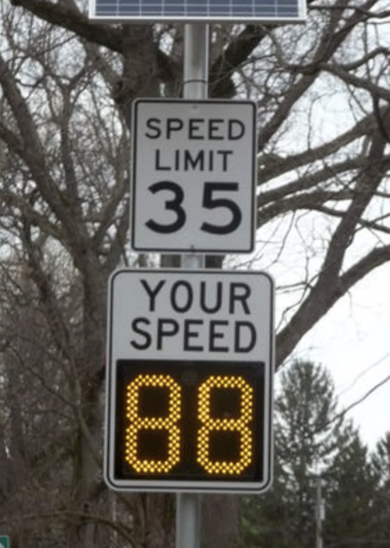 Speed limit sign showing "Speed Limit 35". Below it, an electronic display reads "Your Speed 88". Tree branches in the background