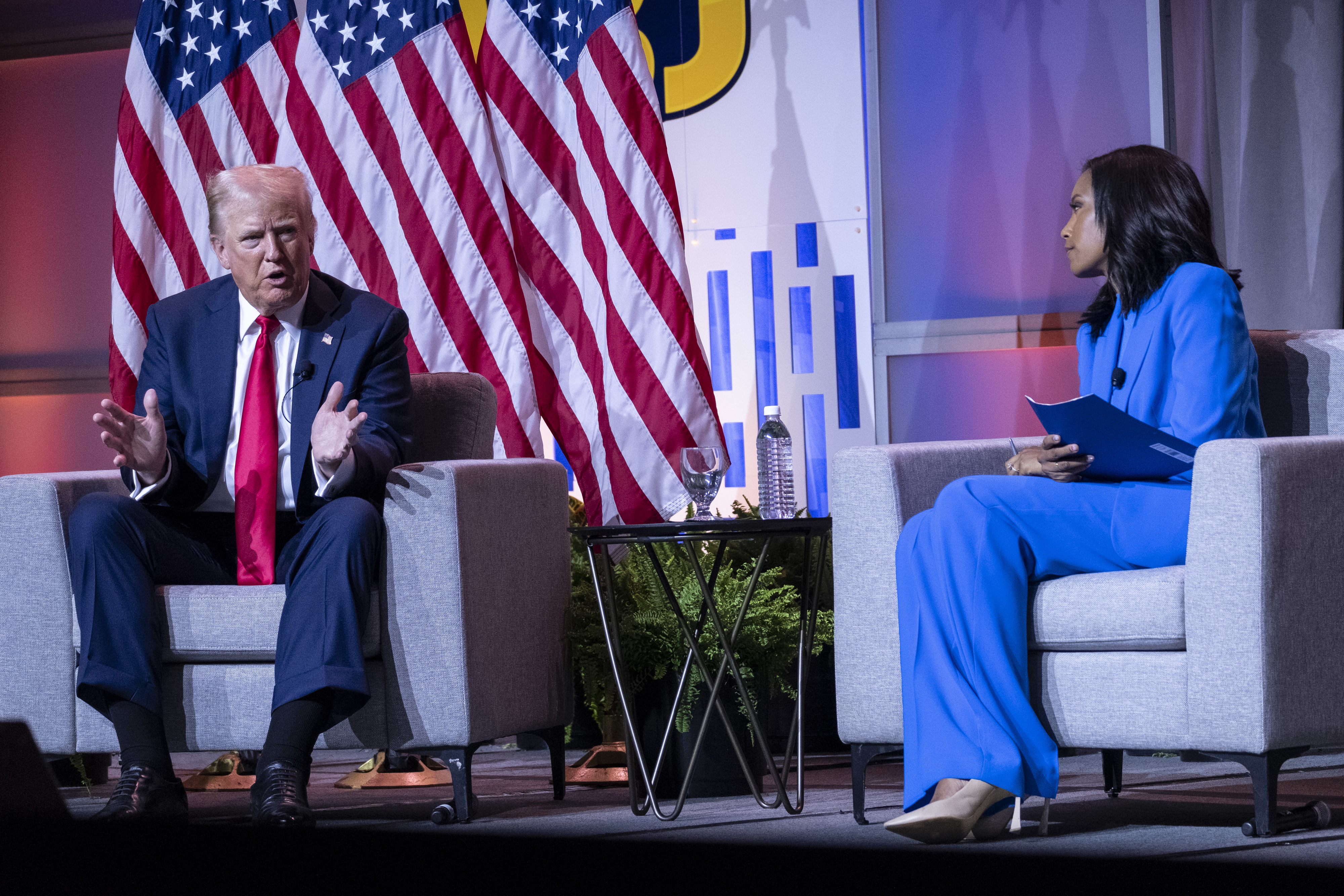Donald Trump speaks on stage, gesturing with his hands, while sitting across from a woman in a blue suit holding documents. Two American flags are in the background