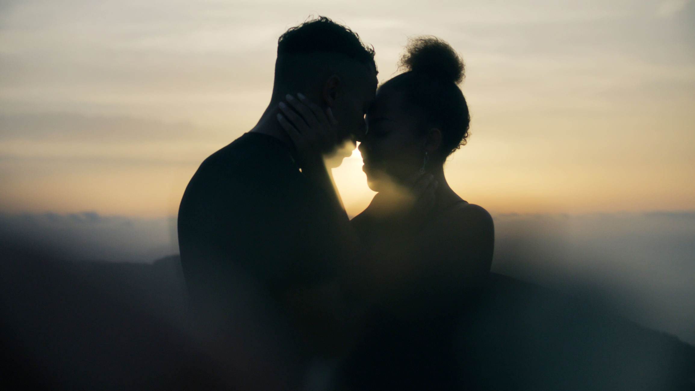 A couple embracing closely with foreheads touching, silhouetted against a sunset backdrop, creating an intimate and romantic scene
