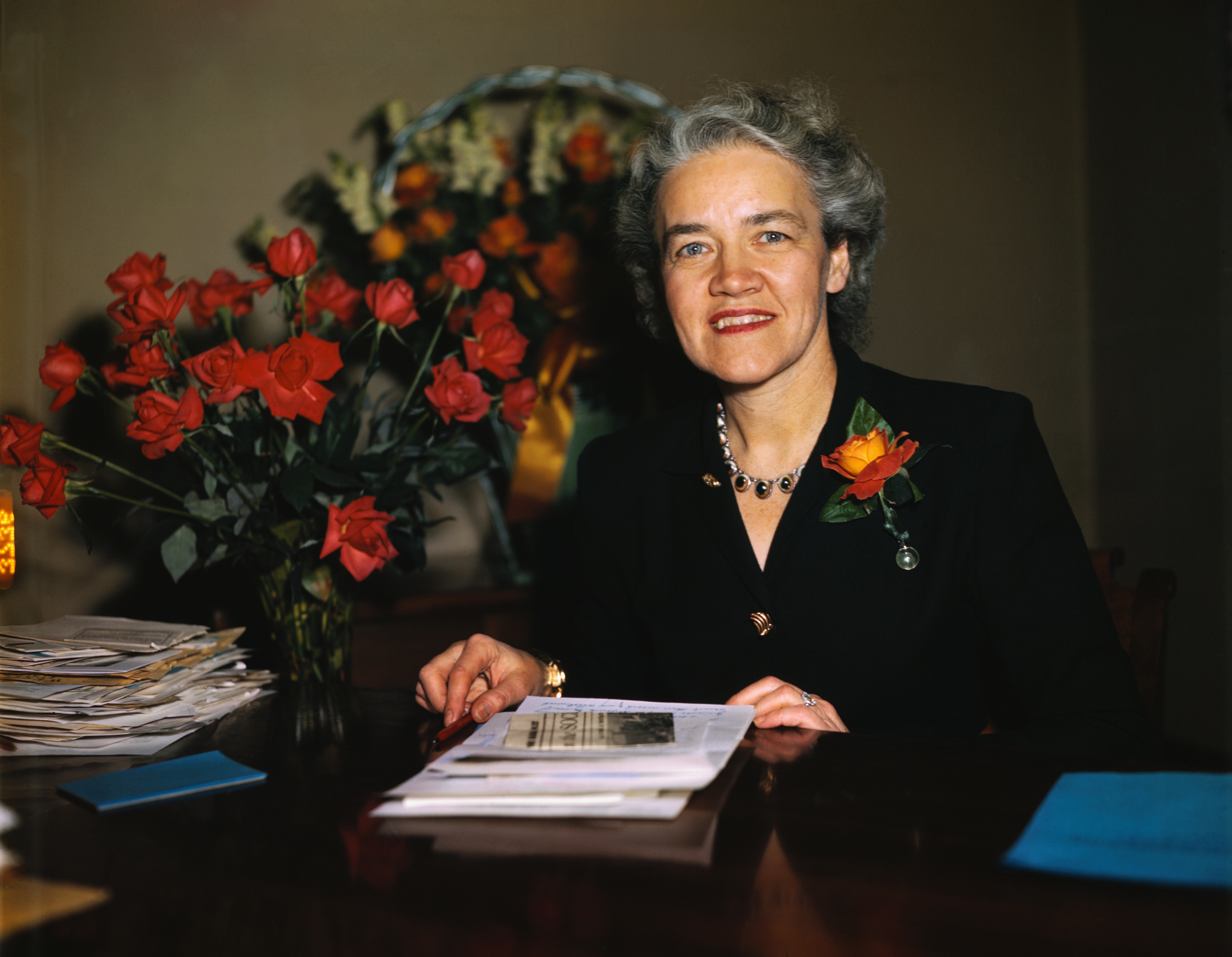 An unidentified person sits at a desk with red roses. They are wearing a dark outfit with a floral brooch and a pearl necklace