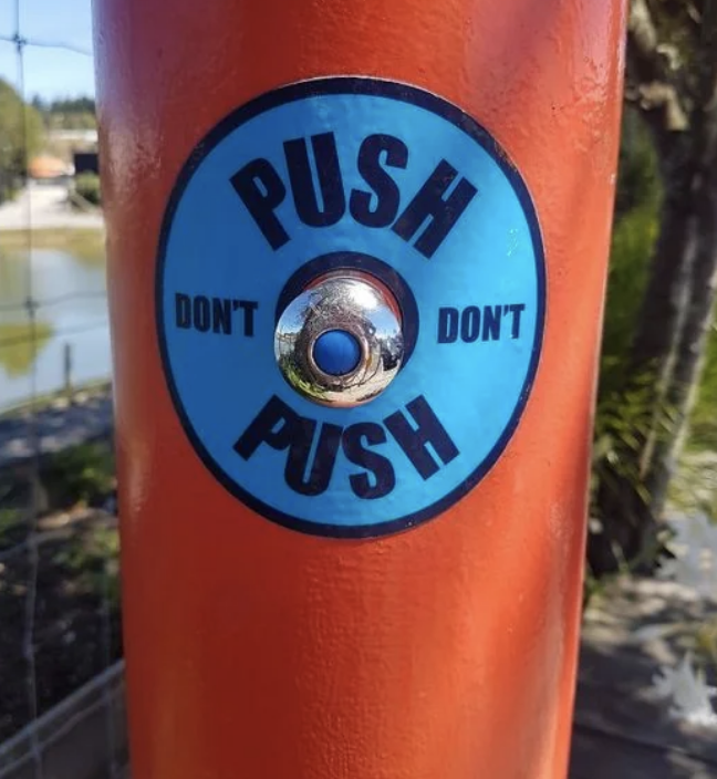 Metal button on an orange pole has conflicting instructions: "PUSH" in large font, and "DON'T" in smaller font twice above and below the button