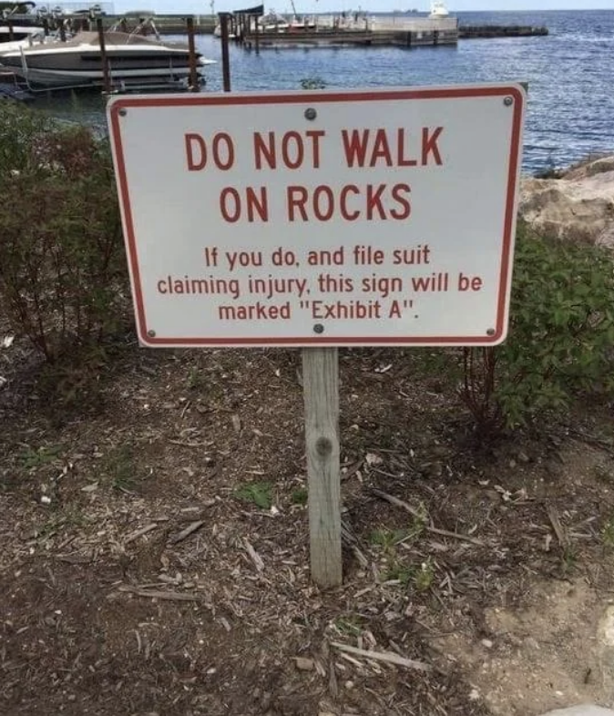 A warning sign near water reads: "DO NOT WALK ON ROCKS. If you do, and file suit claiming injury, this sign will be marked 'Exhibit A'."