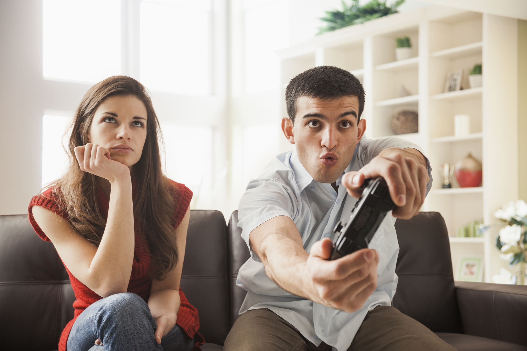 A man excitedly playing a video game sits next to a woman who appears bored and disinterested