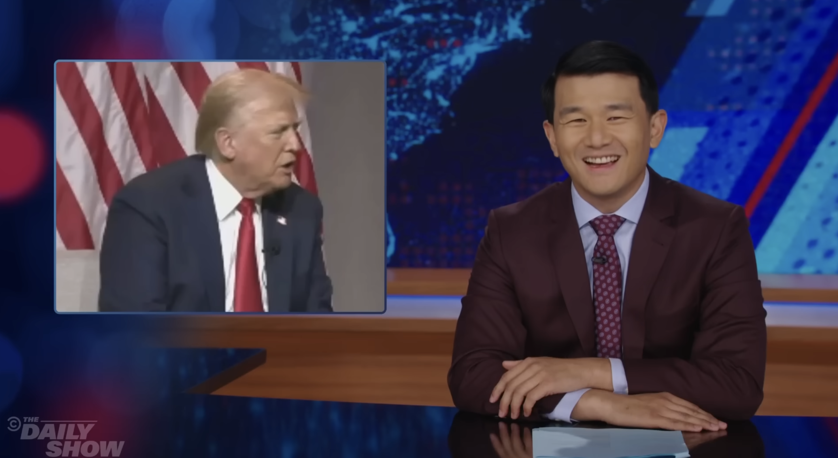 Ronny Chieng, in a suit and tie, hosts The Daily Show with an image of Donald Trump speaking in the background