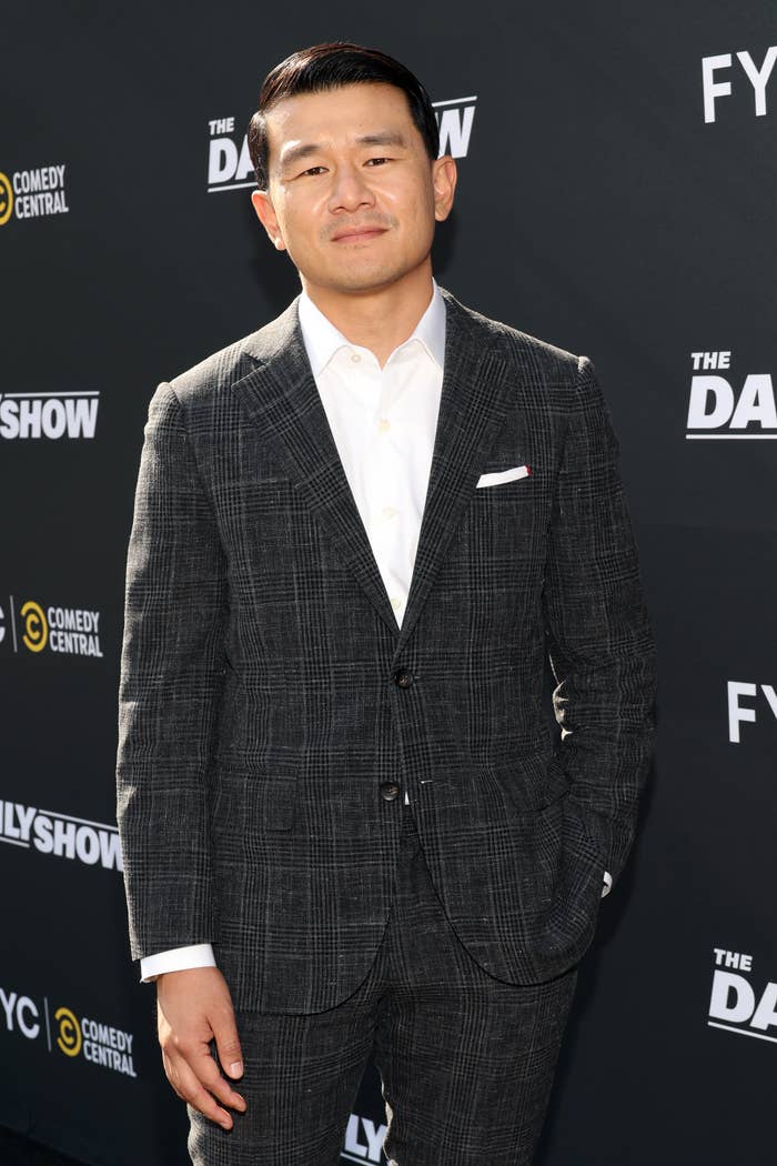 Ronny Chieng stands on the red carpet in a plaid suit with hands in pockets, at The Daily Show event