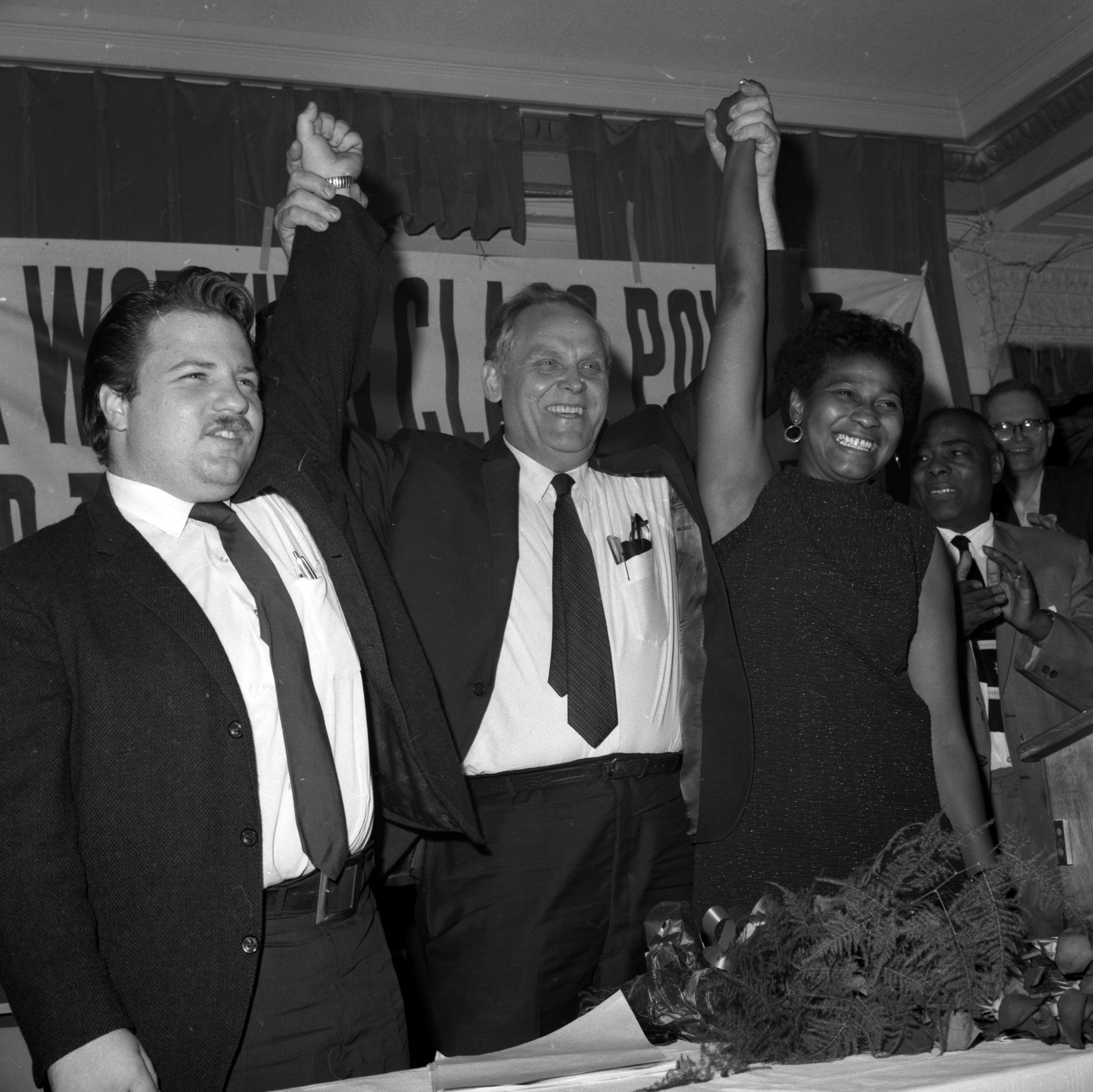People raising arms in victory: Cesar Chavez, George Meany, and an unidentified woman, at an event with a "Support Worker Power" banner in the background