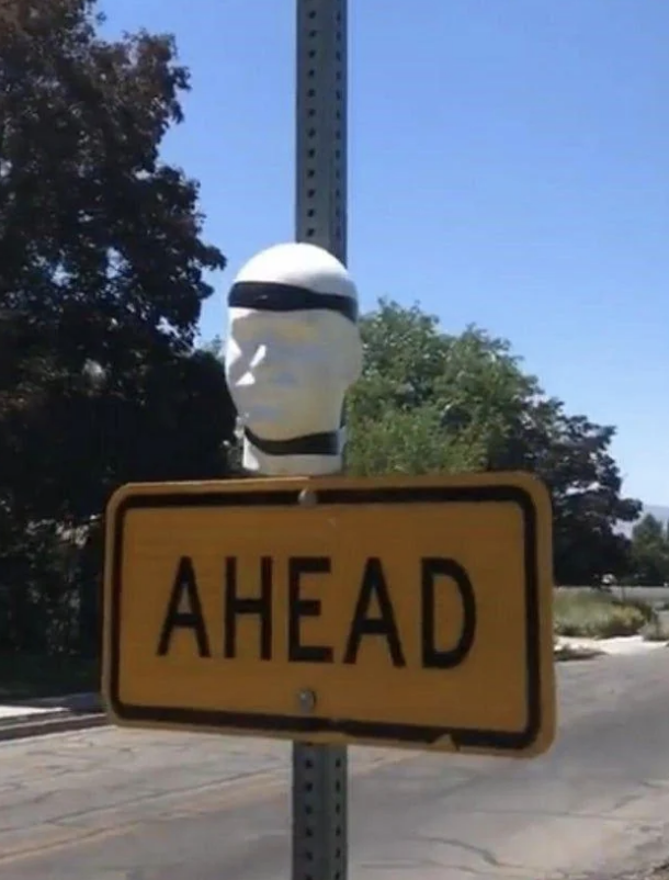 A sign reads "AHEAD" with a mannequin head above it on a pole. Trees and a road are in the background