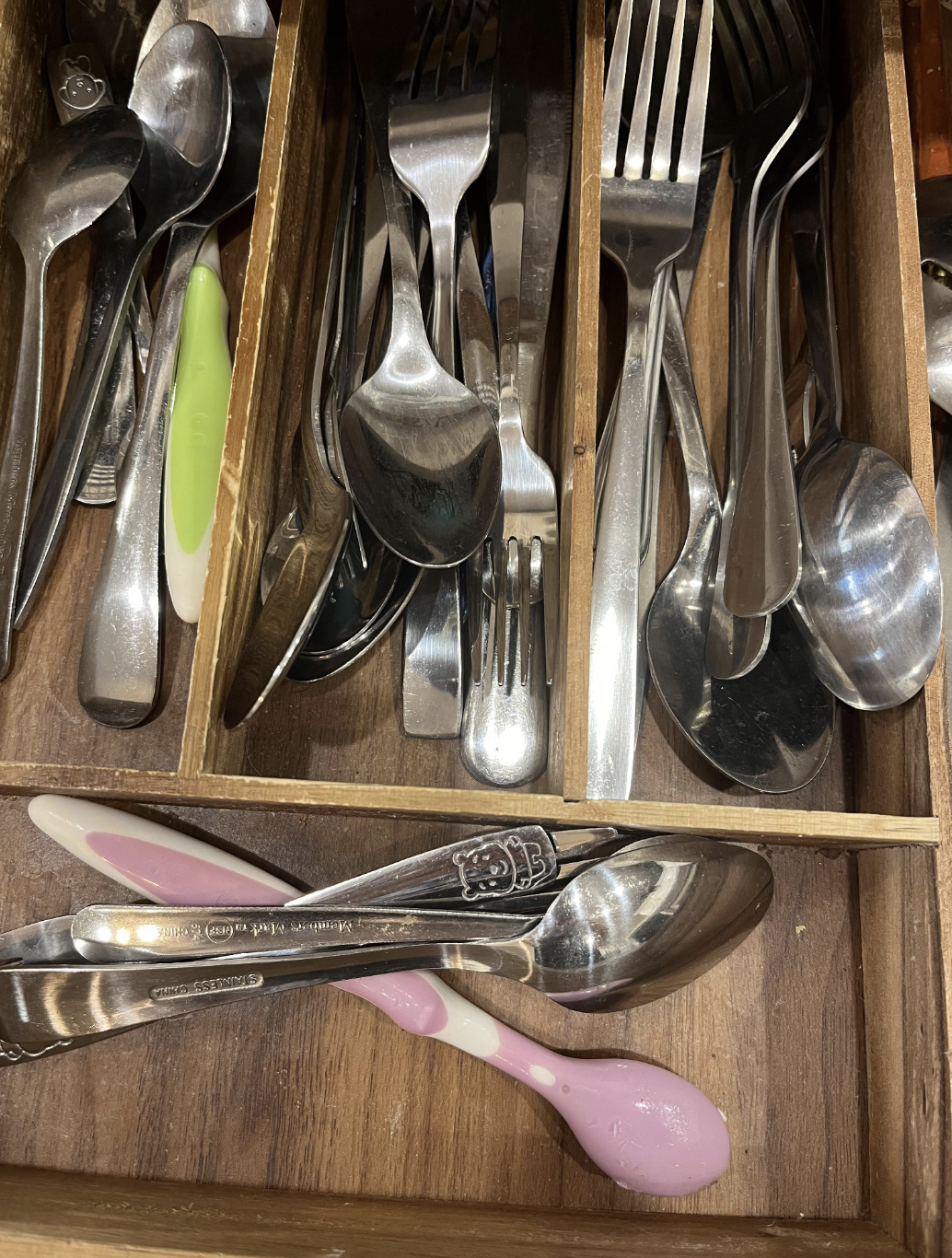 A wooden cutlery drawer organizer filled with various forks, spoons, butter knives, a green-handled spoon, a white and pink spoon, and a silver measuring spoon