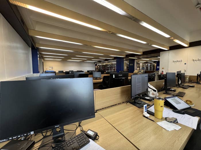 Empty office space with multiple workstations, computers, papers, and personal items on desks. Background shows shelves with books