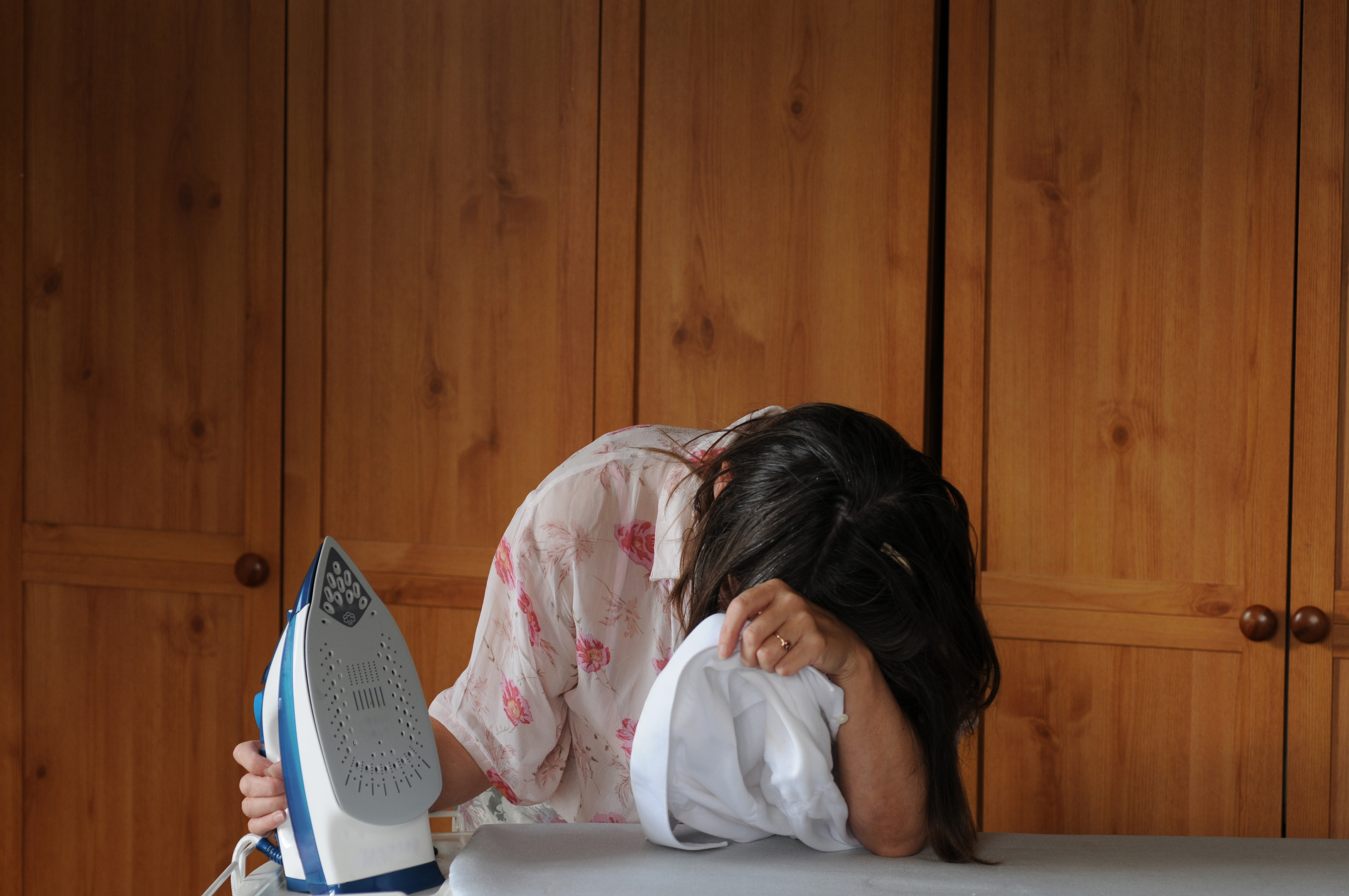 A person with their head in their hand sitting at an ironing board beside an iron, appearing frustrated. Wooden cabinets in the background.