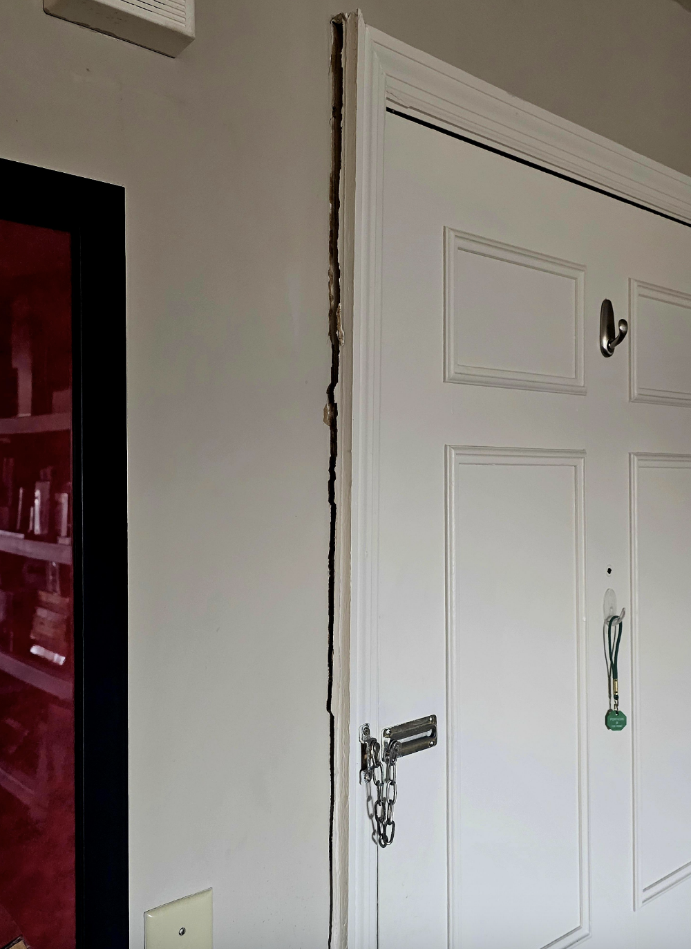 A door with a chain lock is shown. The wall next to the door is damaged, revealing cracks and peeling paint. A light switch is visible to the left
