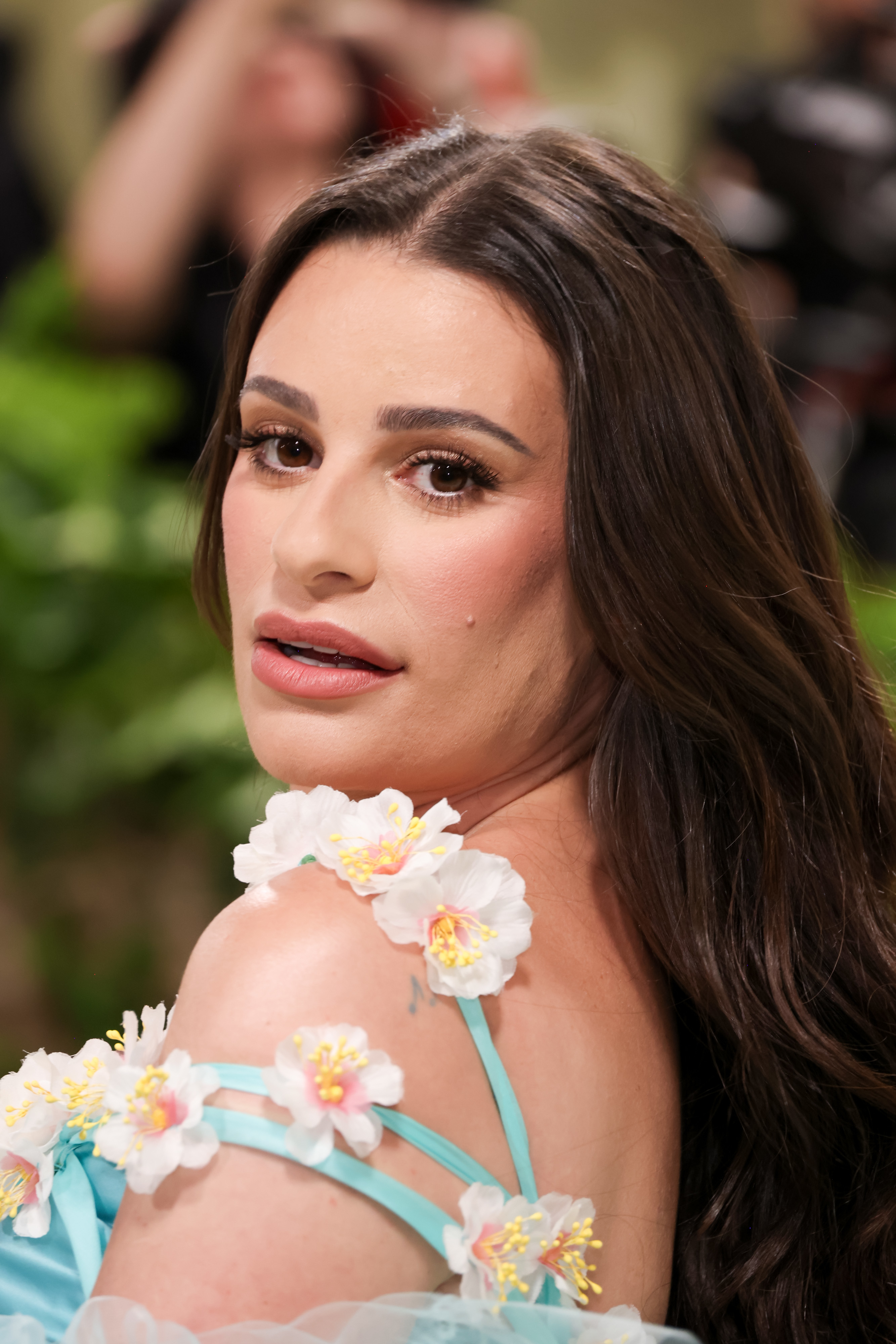 Lea Michele looking over her shoulder, wearing a floral-themed dress with decorative white and pink flowers on the straps and neckline