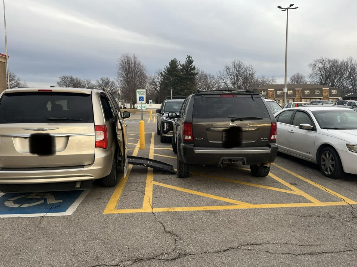 Two SUVs are obstructing a handicap-accessible parking space, with one vehicle partially blocking the ramp, in a parking lot