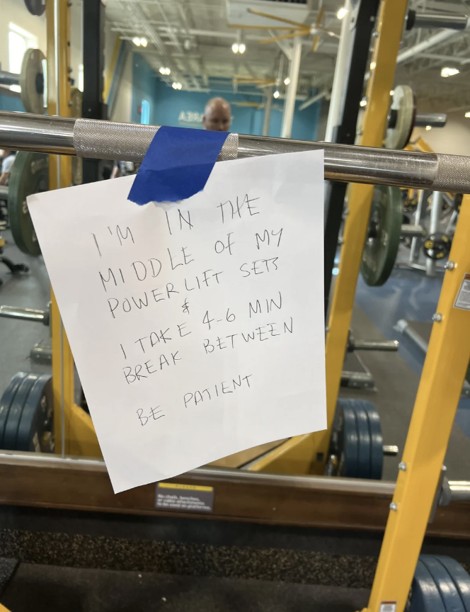 Note on weightlifting bar reads, "I'm in the middle of my power lift sets. I take 4-6 min break between. Be patient." Gym equipment in the background