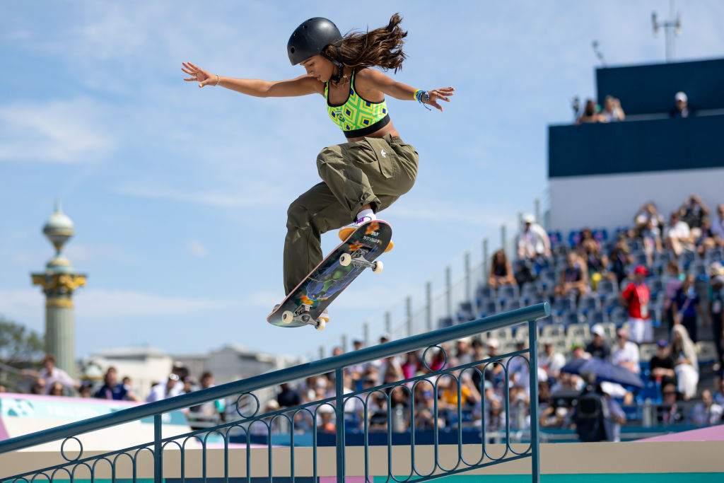 Woman skateboarding down a rail in front of a crowd, wearing a helmet and a crop top with baggy pants
