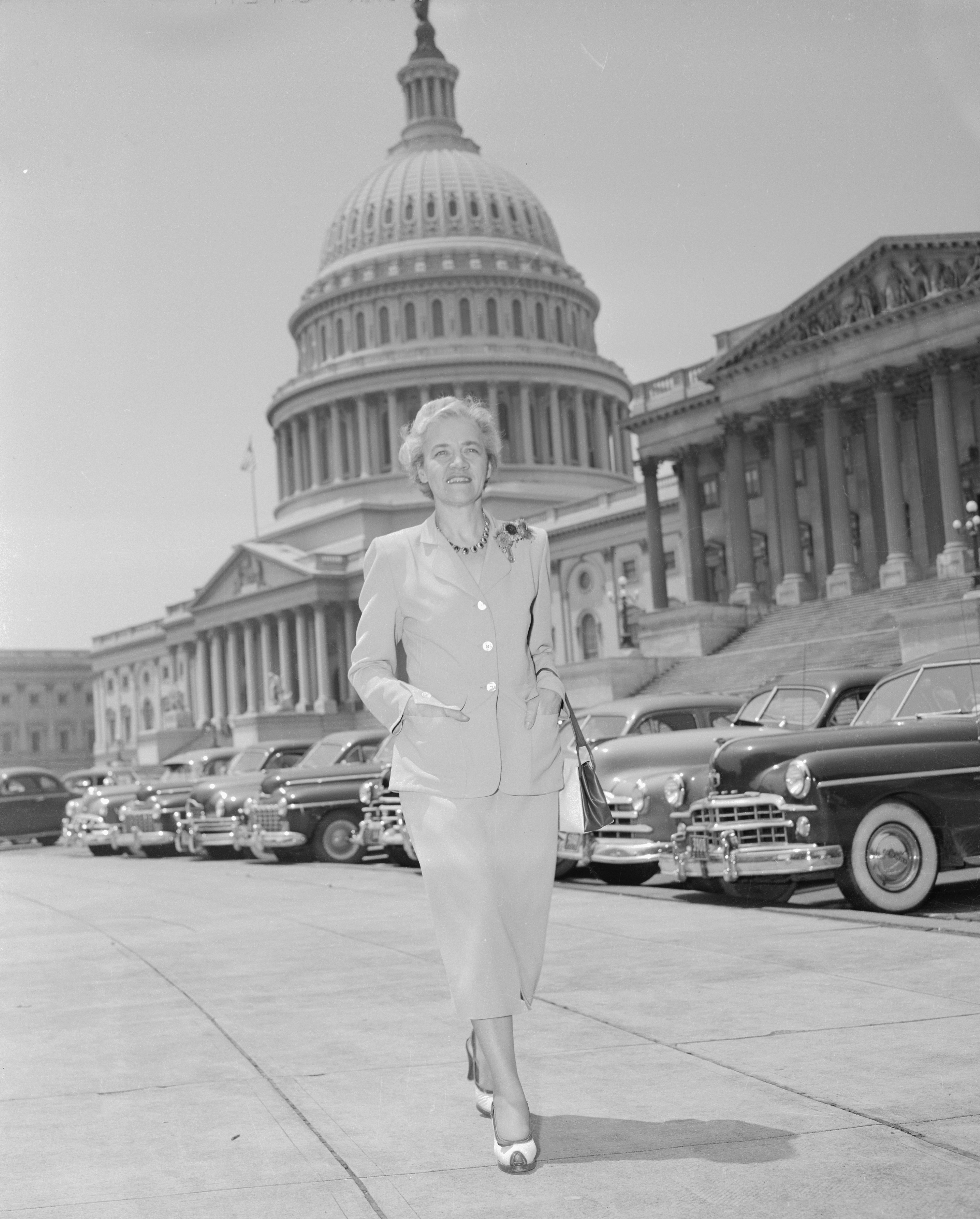 Margaret Chase Smith walks confidently in front of the U.S. Capitol building, wearing a formal suit and holding documents. Classic cars are parked behind her