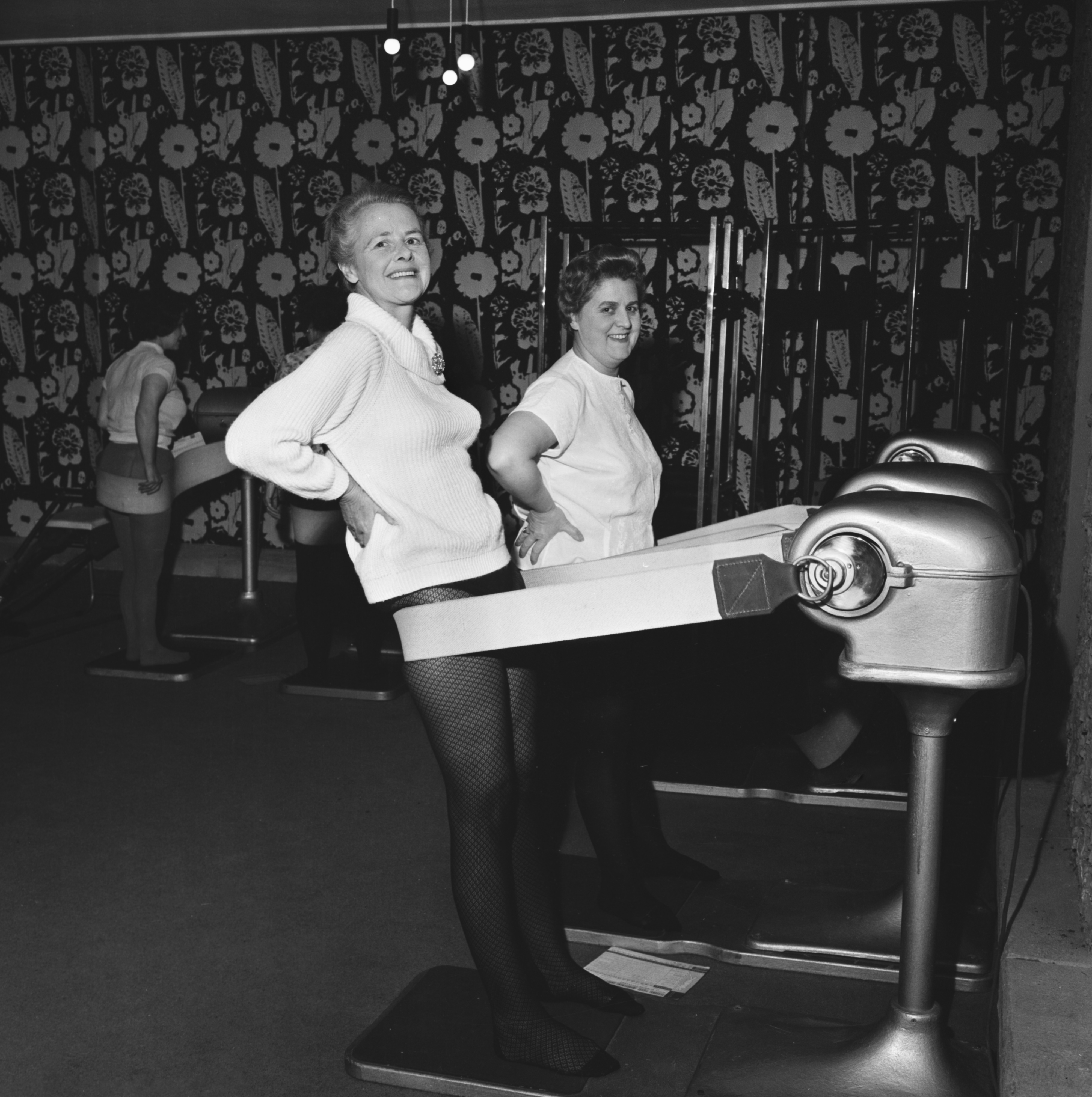 Two women, dressed in casual early 20th-century attire, smiling and using vintage vibration belt exercise machines in a retro fitness center.