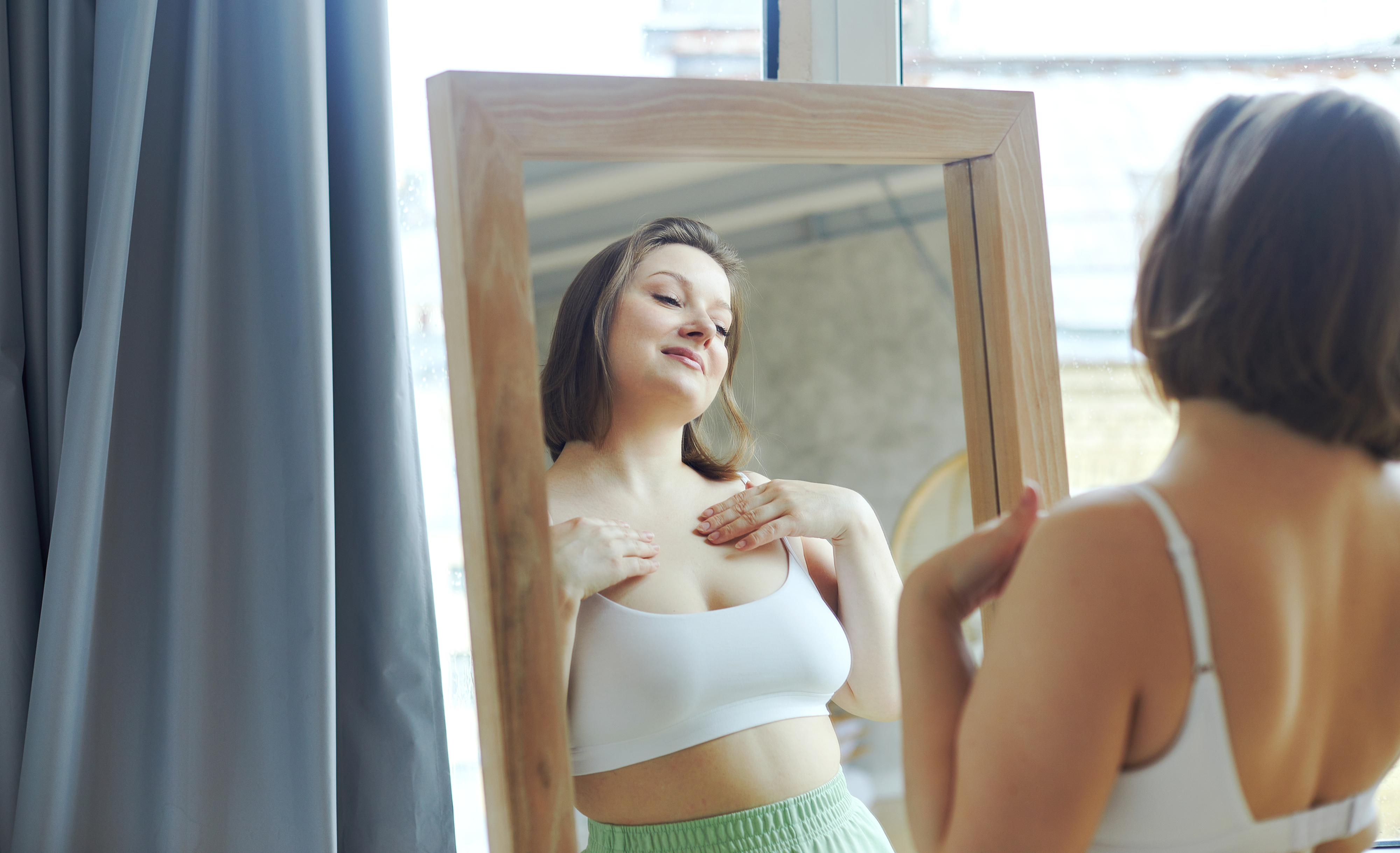 A woman in a white sports bra and green pants looks at herself in the mirror, appearing confident.
