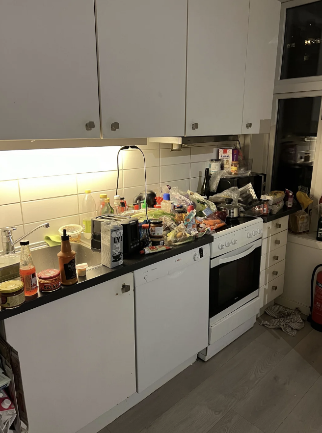 A cluttered kitchen with white cabinets and appliances. The countertop is filled with various food items, bottles, and utensils. There are no people in the image