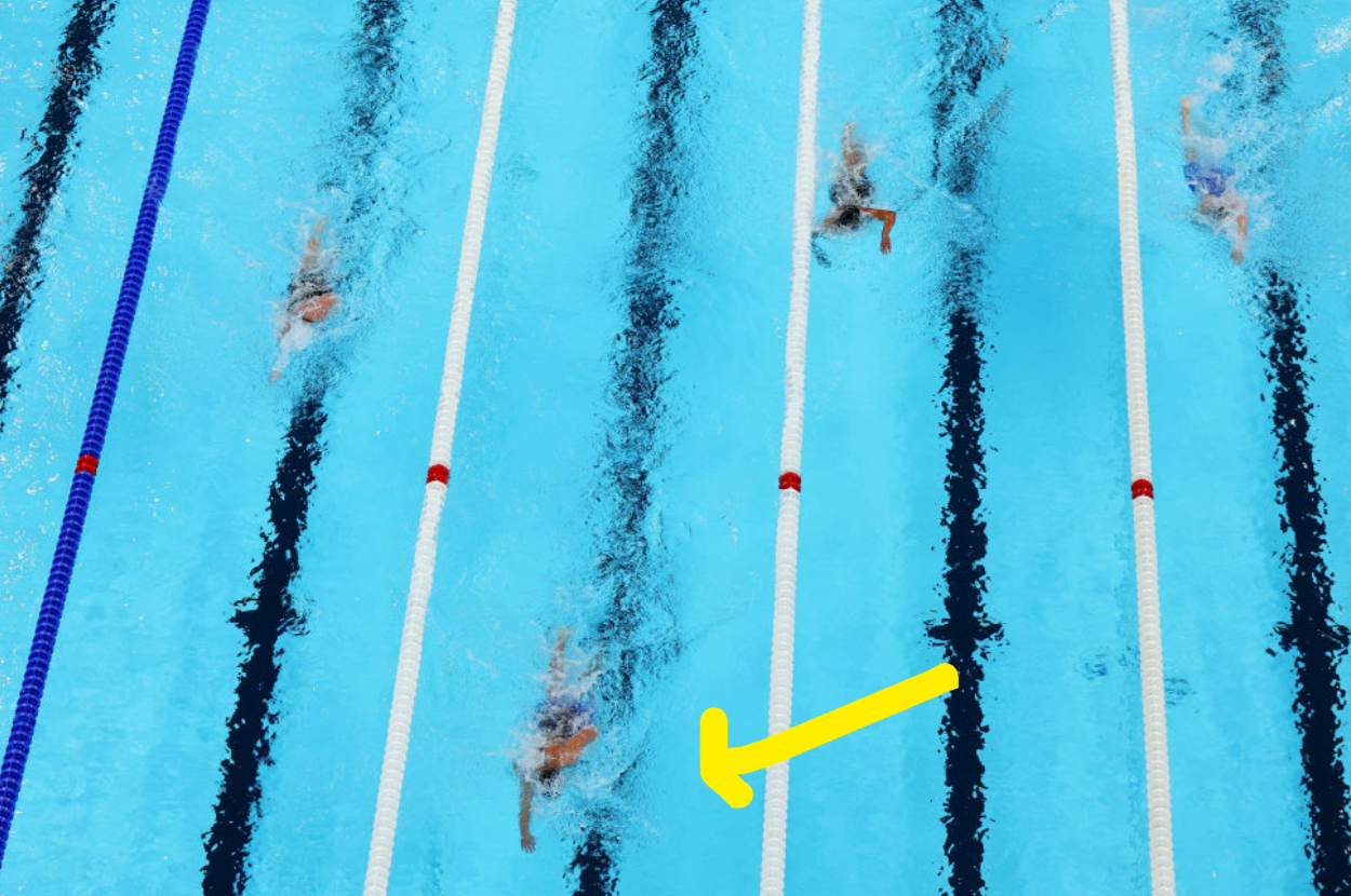 Swimmers in a competitive race are captured mid-stroke in pool lanes, with one swimmer marked by a yellow arrow
