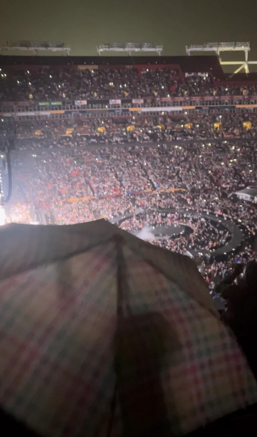 A large, packed stadium at night with a crowd of people watching a concert. A faint image of a stage and lights is visible in the distance. An umbrella in the foreground