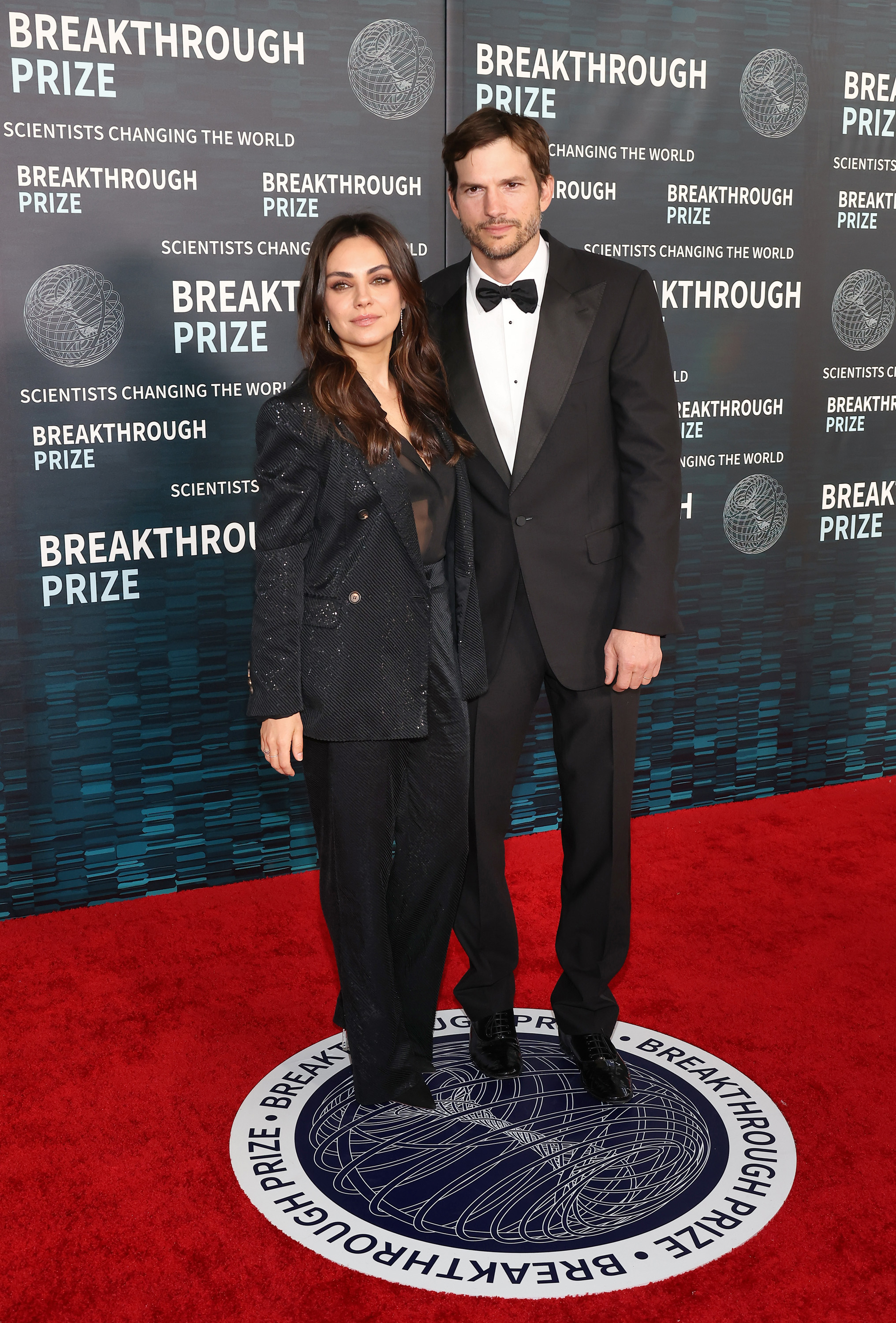 Mila Kunis and Ashton Kutcher on the red carpet at the Breakthrough Prize event. Mila wears a stylish black pantsuit, while Ashton is in a classic black tuxedo