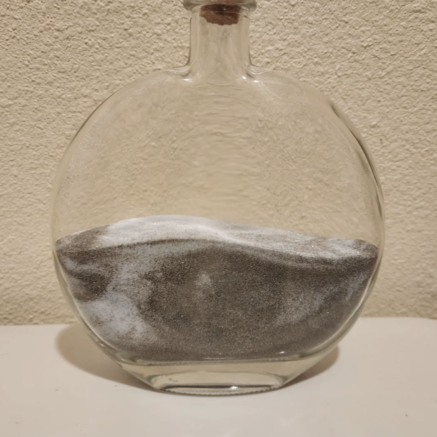 A clear round glass bottle filled halfway with layers of black and white sand, placed against a textured wall