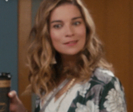 Annie Murphy holding a coffee cup and smiling, wearing a floral-patterned top in a scene from Schitt's Creek. Text at the top reads &quot;#SchittsCreek.&quot;
