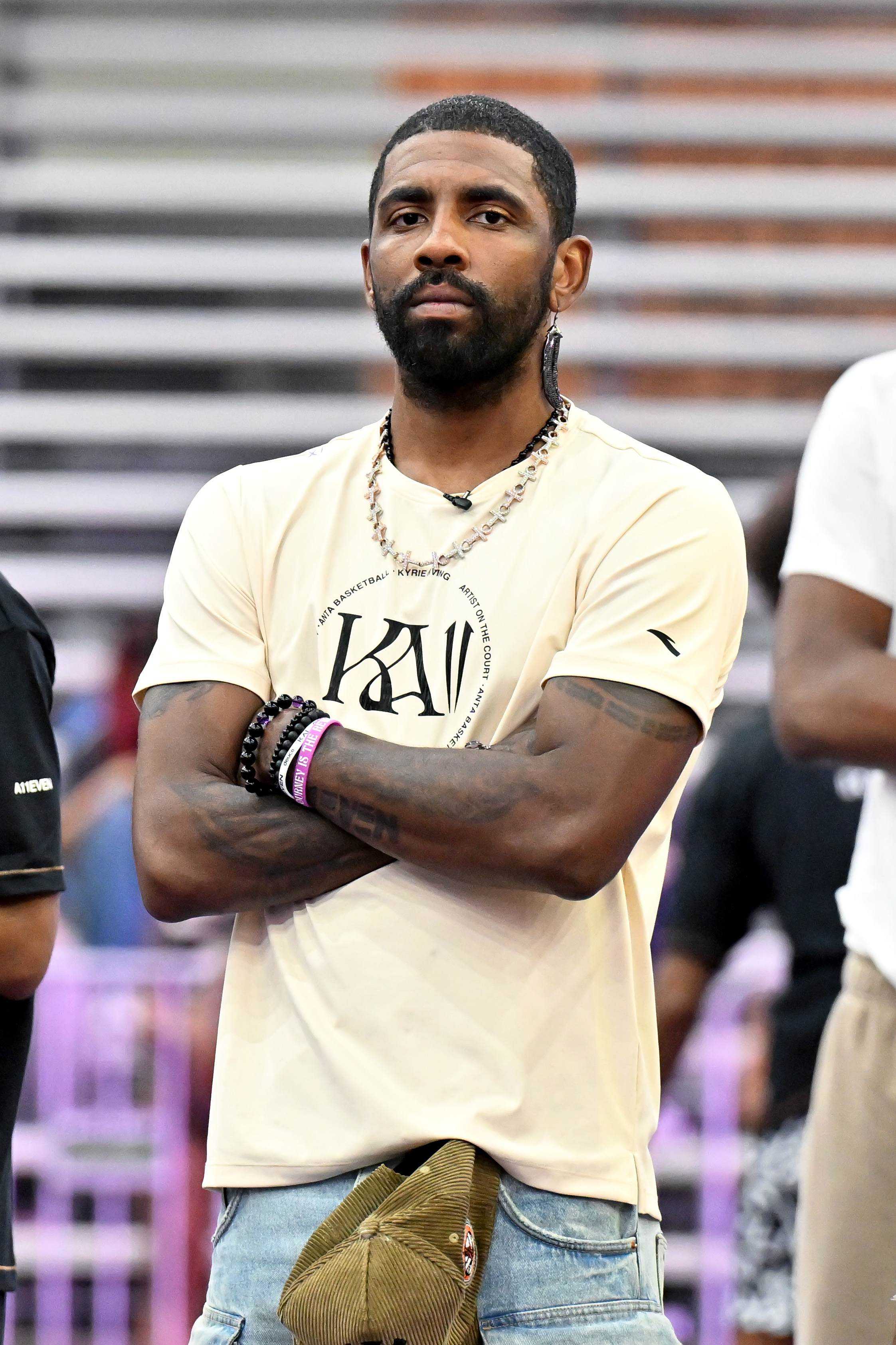 Kyrie Irving stands with arms crossed, wearing a casual t-shirt with a graphic, denim pants, and a necklace