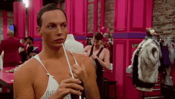 A person wearing a white tank top with thin straps is in a dressing room, applying makeup while looking into the distance