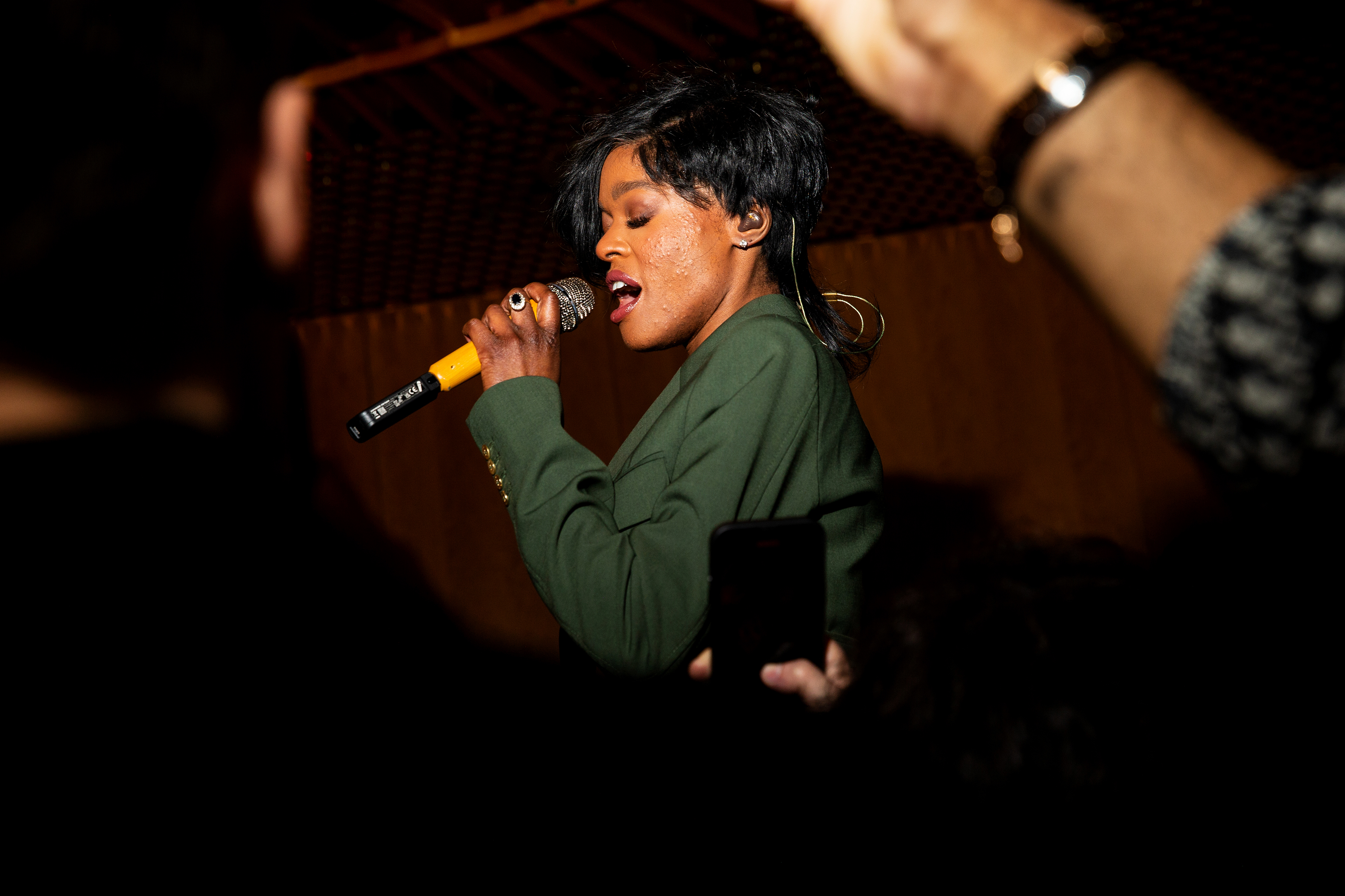 Azalea Banks in a green jacket sings passionately into a microphone on stage. Audience members' arms can be seen in the foreground