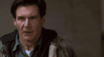 Harrison Ford, in an intense scene from a movie, walks inside a dimly lit location, wearing a coat over a shirt and hoodie