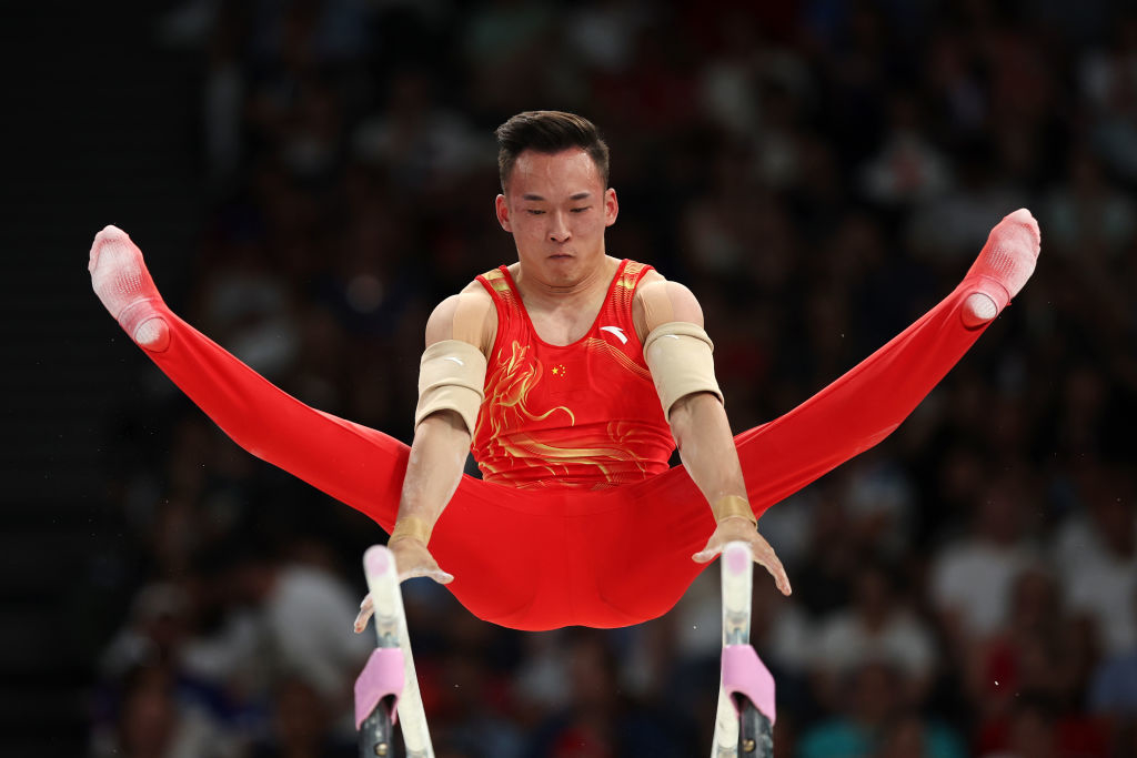 Gymnast performing a split on parallel bars during a competition, wearing a red outfit with gold accents