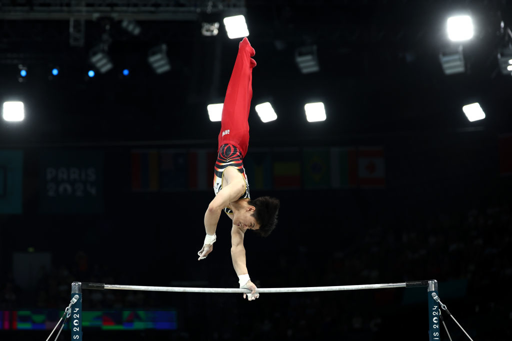 A gymnast performs a handstand on the horizontal bar during a competition at the 2024 Paris event. The background shows blurred flags and lighting