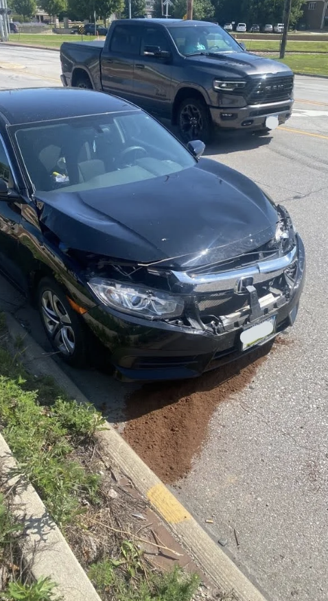 A black sedan with significant front-end damage is parked on the roadside, possibly due to an accident. A black pickup truck is in the background